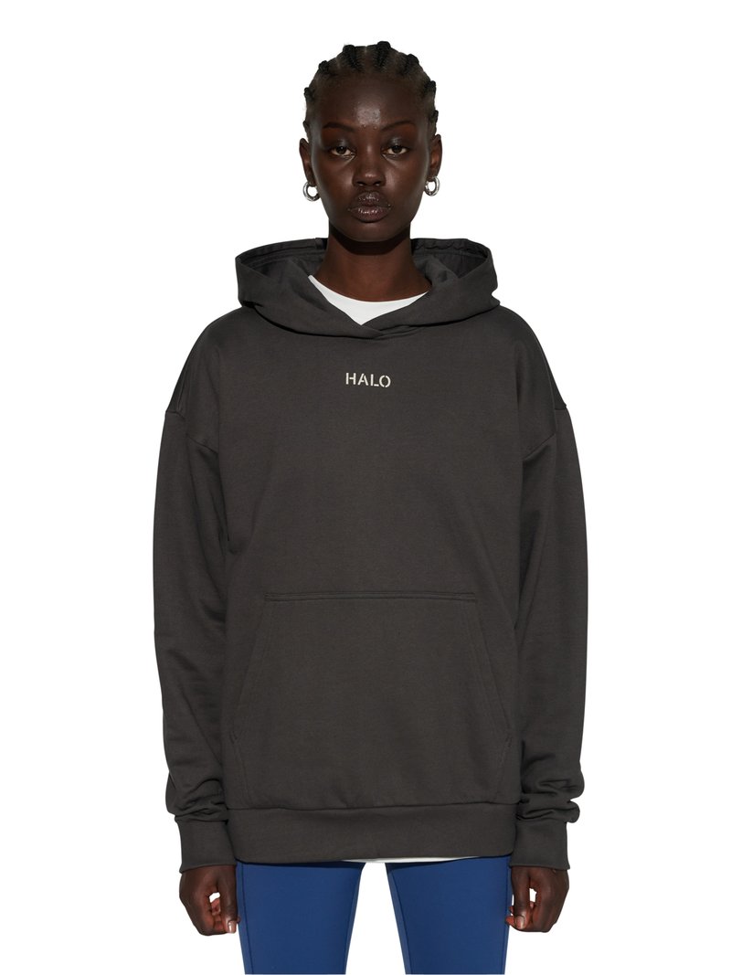 Person trägt schwarzen, übergroßen Hoodie mit dem Schriftzug "HALO" auf der Brust, blaue Hose und silberne Creolen, schaut nach vorne mit neutralem Gesichtsausdruck.