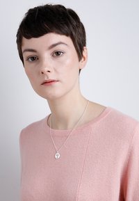 Collier en argent avec un pendentif rond présentant un design d'arbre, porté sur un pull rose pâle à col rond. Texture lisse et style minimaliste.