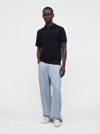 Homme debout portant un polo noir à manches courtes, un jean bleu clair à jambes larges et des baskets blanches sur un fond blanc uni.