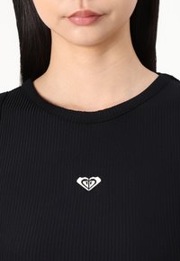 Svart ribbestrikket topp med rund hals og liten hvit hjerte-logo i midten. Materialet ser glatt og litt elastisk ut.