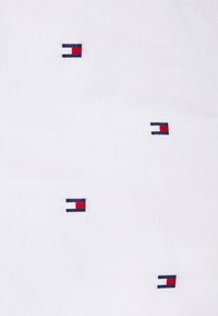 Vitt tyg med flera små broderade loggor i blått, rött och vitt. Loggorna är jämnt fördelade och har en rektangulär design.