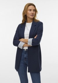 Mornarski blejzer s prilagojenim dizajnom, ki ima enojno gumbovanje, stranske žepe in črtaste manšete. V kombinaciji z belim srajcem in kavbojkami.