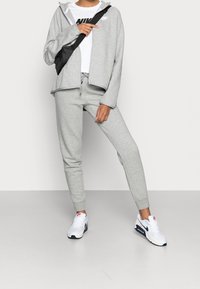 Ljusgrå hoodie med dragkedja och sidofickor, matchad med sweatpants. Tillbehör inkluderar en svart axelväska och vita sneakers.