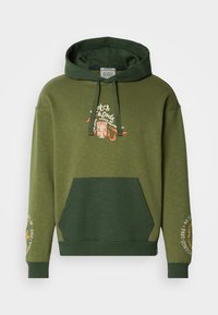 Ένα ελαιόχρωμο φούτερ με καζάκα που διαθέτει τσέπη καγκουρό, κουκούλα με κορδόνι και κεντημένες λεπτομέρειες στα μανίκια για υφή και σχέδιο με πολλές στρώσεις.