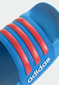 Niebieska sandałka na platformie z trzema czerwonymi, paskowanymi akcentami. Teksturowana gumowa powierzchnia i okrągłe białe logo na boku.