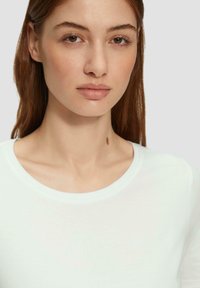 Chemise blanche à manches courtes avec un col rond, fabriquée en matériau léger. Le mannequin a les cheveux bruns raides et une petite grain de beauté sur le cou.