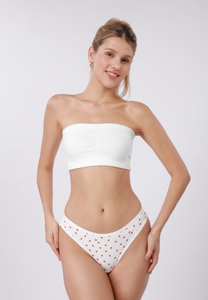 Νεαρή γυναίκα που φορά λευκή τοπ χωρίς τιράντες και λευκό μπικίνι με μικρές κόκκινες καρδιές, στέκεται μπροστά σε απλό φωτεινό φόντο, χαμογελώντας.