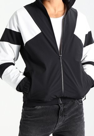 Mies pukeutuneena mustavalkoiseen vetoketjulliseen takkiin, kädet taskuissa, yhdistettynä tummanharmaisiin farkkuihin, yksinkertaisella taustalla.