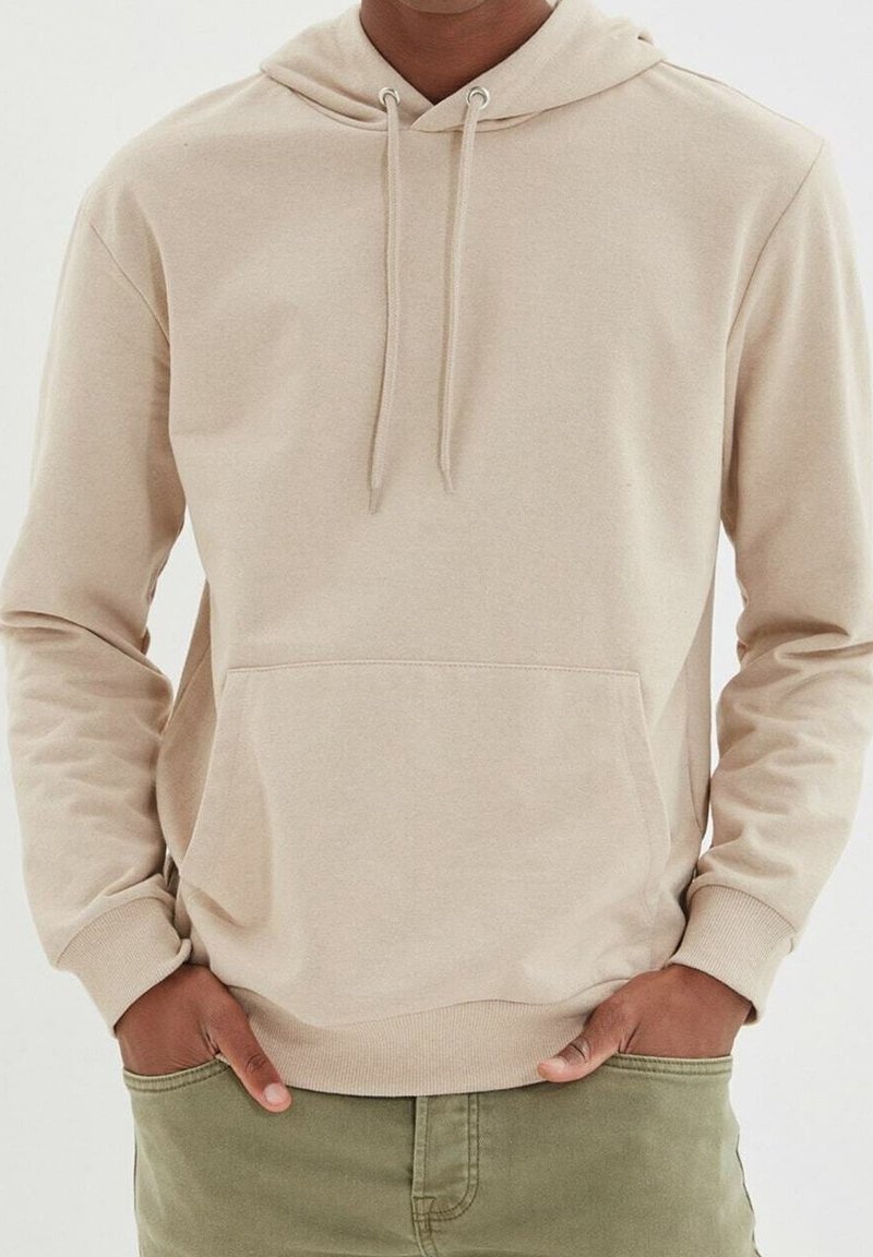 Uomo che indossa una felpa con cappuccio beige con tasca frontale e pantaloni verdi, mani nelle tasche, in piedi contro uno sfondo bianco.