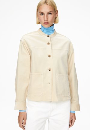 Woman with short blonde hair wearing a beige buttoned jacket over a bright blue turtleneck and white pants, standing against white background.