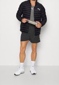 Černá péřová bunda, šedé sportovní tričko, tmavé šortky, bílé sportovní boty, držící stříbrnou láhev na vodu. Logo viditelné na oblečení a botách.