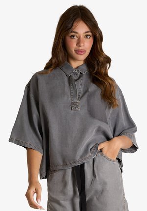 Donna con capelli lunghi castani che indossa una maglietta grigia oversize a maniche corte e pantaloni larghi abbinati con la mano in tasca.
