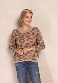 Beiger Pullover mit braunem abstraktem Muster, rundem Ausschnitt und gerippten Bündchen. Kombiniert mit zerrissenen blauen Jeans. Weiche Textur und lockere Passform.