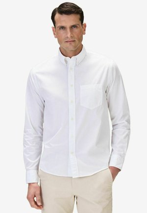 Mand med kort mørkt hår iført en hvid skjorte med knapper og lomme samt beige bukser, stående med den ene hånd i lommen.