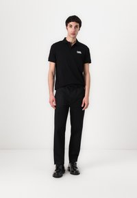 Čierne polo tričko s krátkymi rukávmi a značkovým logom na hrudi, v kombinácii s čiernymi nohavicami a čiernymi topánkami. Jednoduché pozadie.