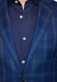 Granatowa marynarka w kratę o fakturowanej powierzchni, zapinana na pojedynczy guzik, oraz pasująca ciemna koszula z subtelnym splotem i jasnymi guzikami.