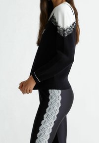 Pull noir avec des détails en dentelle blanche sur les épaules, associé à des leggings noirs ornés d'une bordure en dentelle blanche sur les côtés, manches longues.