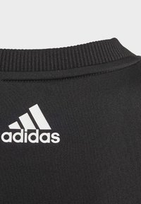 Černý svetr s límcem, který má ribbed strukturu a výrazné bílé logo Adidas na zadní straně. Hladká tkanina bez dalších akcentů.
