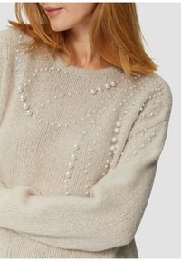 Frau mit rotem Haar, die einen beigen Strickpullover trägt, der an der Vorderseite und an den Ärmeln mit verschiedenen Größen von aufgenähten weißen Perlen geschmückt ist.