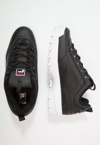 Schwarze Ledersportschuhe mit weißer Gummisohle, ausgestattet mit einer strukturierten Oberkante, Perforationen und Logo-Akzenten an der Zunge und Ferse.