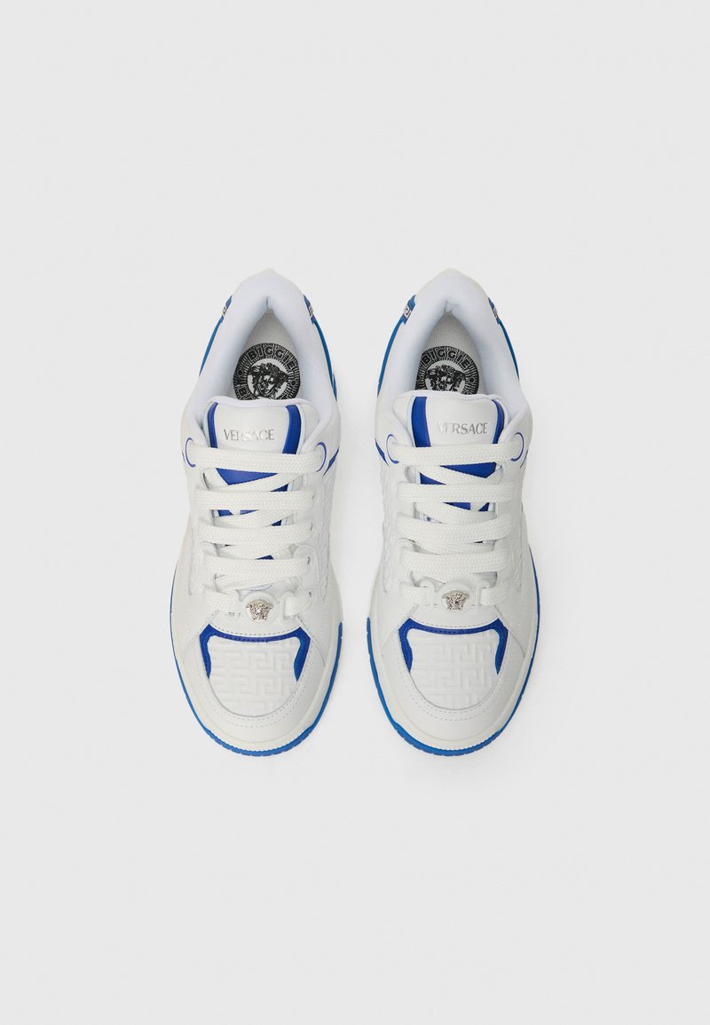 Baskets blanches avec des accents bleus, présentant un panneau latéral texturé, un système de laçage et le logo Versace sur la languette et la semelle.