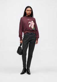 Sudadera burdeos con bordado floral, combinada con jeans negros de cintura alta y botas de tobillo; modelo sosteniendo un bolso negro.