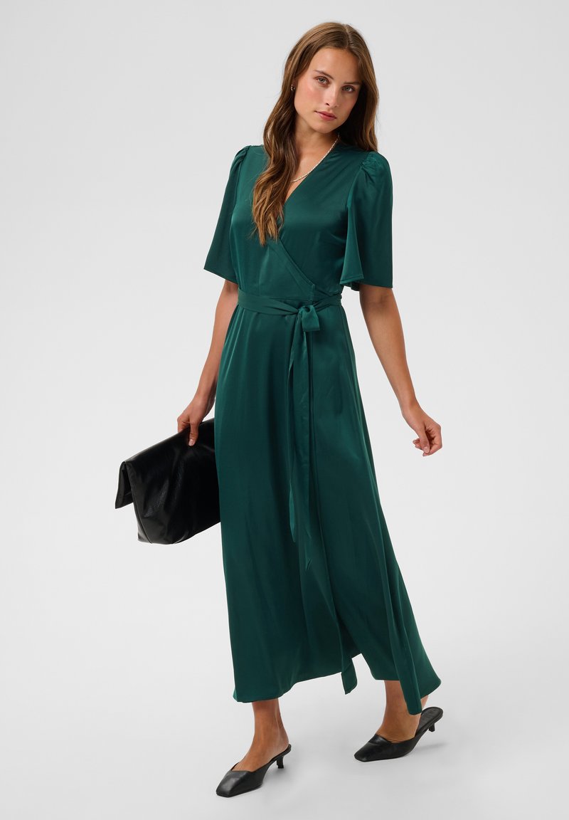 Femme en robe portefeuille vert foncé à manches courtes tenant une pochette noire et portant des chaussures noires à talons bas sur fond uni.