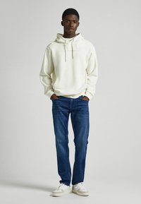 Sudadera blanca con capucha y cordones, bolsillo delantero, combinada con jeans oscuros y zapatillas blancas. El modelo se encuentra de pie contra un fondo claro.
