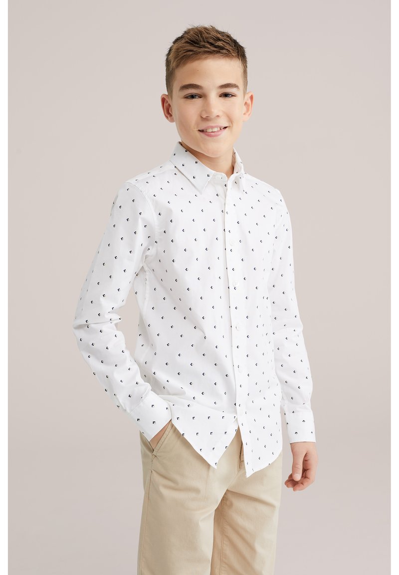 Mosolygó fiú, fehér, apró fekete mintás gombos ingben és bézs nadrágban, egyik kezét a zsebében tartva áll.