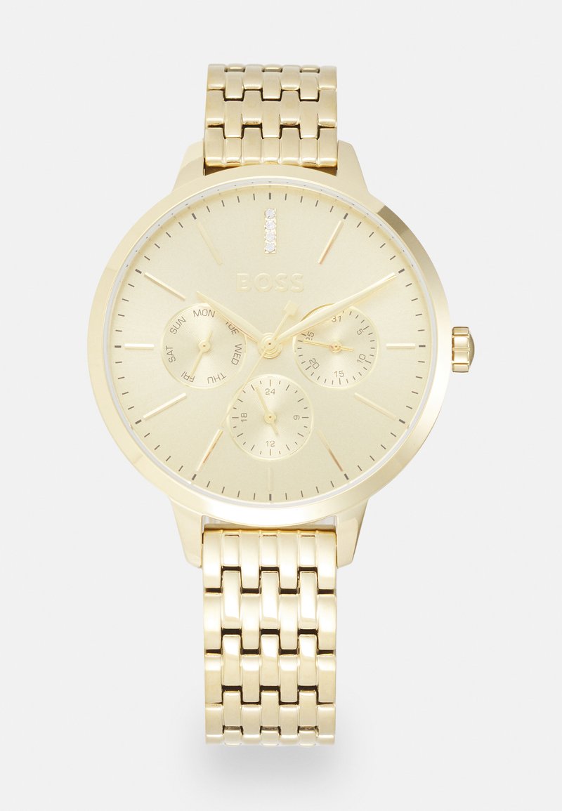 Kuldtooni käekell tekstuurse metallist rihmaga, ümmargune korpus ning minimalistlik dial, millel on kolm aldiali, ning kristallaktsent kell 12.