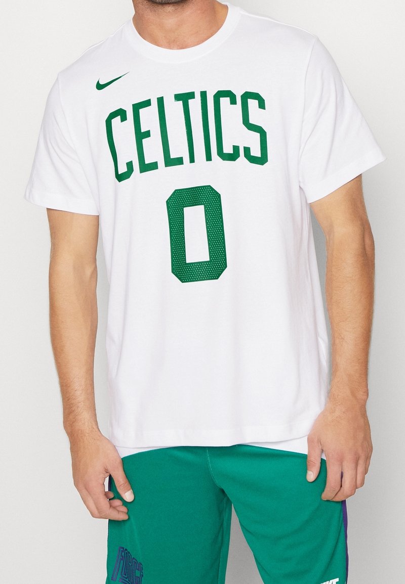 Homme portant un t-shirt blanc des Celtics avec le numéro zéro vert et un short vert, debout devant un fond uni.