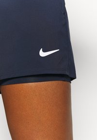 Gros plan sur un short de sport Nike bleu foncé porté sur une cuisse brune, montrant le logo blanc Nike swoosh sur le côté gauche.