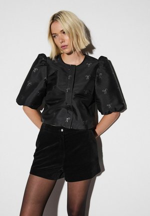 Schwarze Satinbluse mit gepufften Ärmeln, verziert mit silberner Schleifenstickerei und rundem Halsausschnitt. Kombiniert mit schwarzen Shorts und Strumpfhosen.