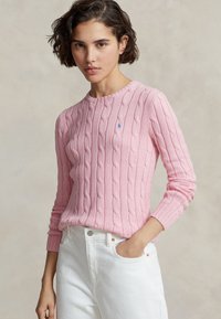 Pull en maille torsadée rose clair avec un col rond et des manches longues. Présente un petit logo sur la poitrine. Porté par-dessus un jean blanc.