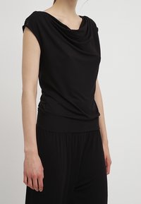 Top negro drapeado con mangas cortas, que presenta un cuello de drapeado y una cintura ajustada. Hecho de una tela lisa y elástica.