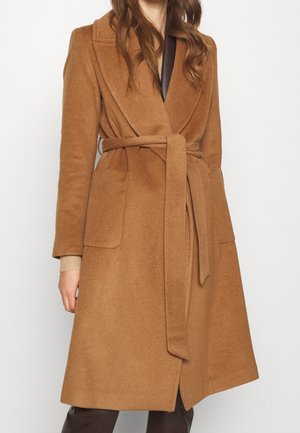 Kobieta w wełnianym płaszczu koloru wielbłądziego brązu z paskiem i dużymi przednimi kieszeniami, ubrana na ciemne spodnie, stoi na tle gładkiej białej ściany.