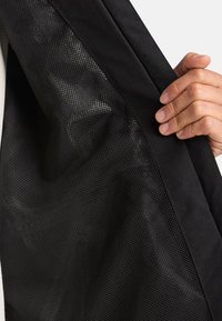 Svart textilväska med nätpanel, som visar ett slätt yttre tyg och en texturerad nätinredning. Innehåller en hand som greppar kanten.