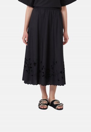 Donna che indossa una gonna midi nera con dettagli floreali traforati e orlo smerlato, abbinata a sandali neri open-toe con fiocchi di corda.