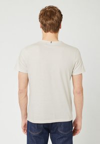 Lichtbeige T-shirt met korte mouwen en een ronde hals, zachte textuur en een klein zwart lusje aan de achterkant. Draag het met donkere denim jeans.
