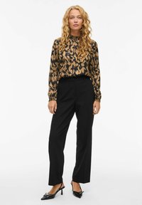 Blusa a maniche lunghe con stampa animale (marrone e nera) abbinata a pantaloni neri su misura. Le scarpe sono nere con punta appuntita e tacco medio.