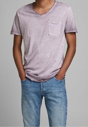 Homme portant un t-shirt violet délavé à col en V avec une poche poitrine et un jean bleu clair, les mains posées négligemment dans les poches.