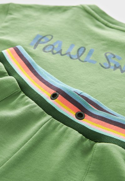 Zaļš kokvilnas apģērbs ar ribotu jostasvietu, kas rotāta ar daudzkrāsainām svītrām un zila skripta logotipu netālu no kakla.