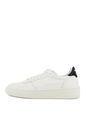 Baskets basses blanches avec semelle crème, fermeture à lacets, détails discrets de couture, et languette noire au talon avec lettrage blanc.