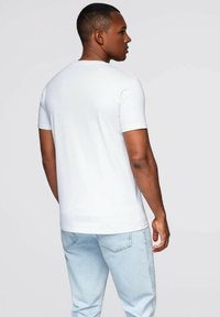 Mand, der står med siden til iført en ensfarvet hvid T-shirt med korte ærmer og lyseblå jeans foran en neutral baggrund.