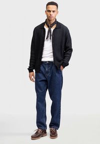 Fekete bordázott, gombos kardigán fehér ing fölött, laza kék farmernadrággal és barna cipővel párosítva, fekete mintás sállal kiegészítve.