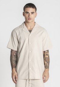 Camisa de botones de manga corta beige con un cuello relajado, hecha de una tela texturizada, que presenta un brillo sutil y cinco botones en la parte delantera.