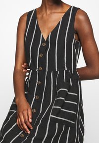 Robe rayée noire et blanche avec un décolleté en V, devant à boutons et poches latérales. Fabriquée en tissu texturé avec un bustier ajusté et une jupe évasée.