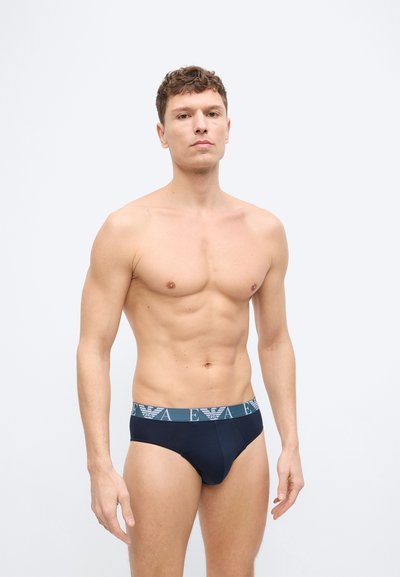 Άνδρας χωρίς πουκάμισο, στέκεται μπροστά σε απλό φόντο, φορώντας σκούρα εσώρουχα με μεταλλικό λογότυπο στη μέση.