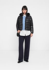 Vrouw in een zwarte pufferjas met een nepbontkraag, blauwe cardigan, wit gestreept shirt, marineblauwe wijde broek, zwarte schoenen en een zwarte schoudertas.