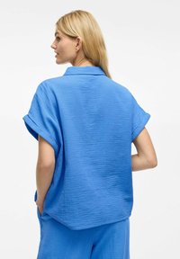 Camicia corta blu con texture, dal taglio morbido e con maniche arrotolate, caratterizzata da un colletto e una tasca sul lato sinistro.